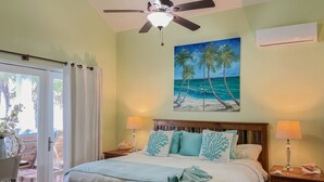 A bedroom with a bed, two bedside lamps, a ceiling fan, a painting of a beach, and a sliding glass door leading to an outdoor area. - Casa Maravilla,Sleeps 8@SapphireBeachResort (Not available)