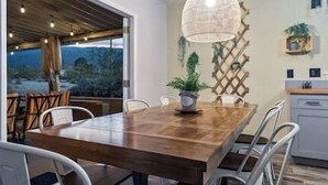 A dining area with a wooden table and white chairs, a hanging light fixture, and a view of the outdoors. - DesertDreamscape Sauna Cold Plunge SPA 3 min to JT (Twentynine Palms)