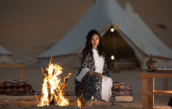 A tent with a person sitting by a fire, surrounded by cushions and a table with a teapot. - Safari Algeria Camp (Djanet)