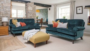 Un salon avec deux canapés de couleur turquoise, un pouf jaune moutarde, un mur en pierre et, au fond, un coin repas.