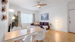 Una sala de estar moderna con un sofá, una mesa de comedor y sillas. Cuenta con un ventilador de techo, una ventana con cortinas y una puerta.