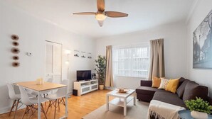 Una sala de estar moderna con comedor, ventilador de techo, televisor y un amplio ventanal con cortinas.