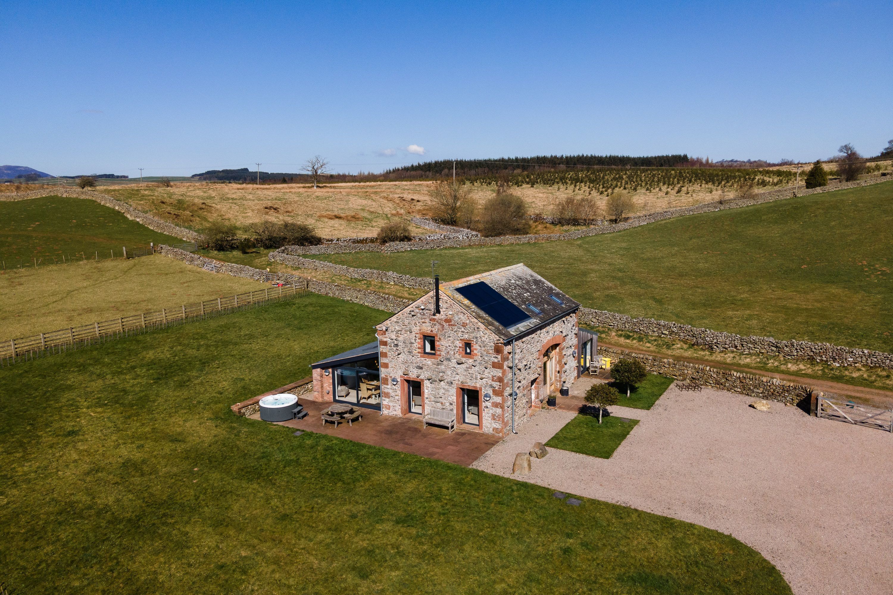 A stone house with a solar panel, surrounded by a stone wall and a grassy field.