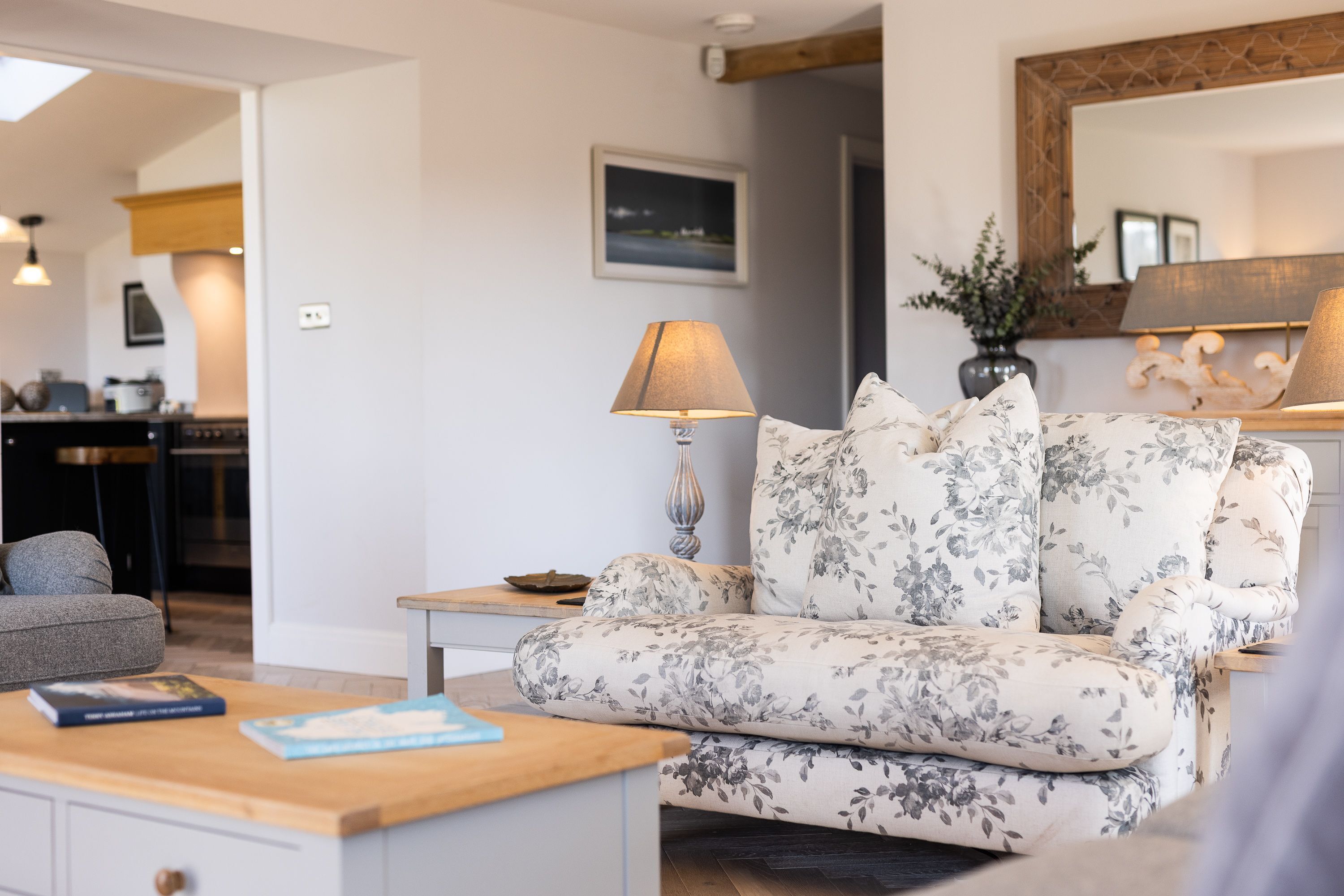 Un salon avec un canapé à motifs floraux, une table basse en bois, une lampe et, au fond, une cuisine.