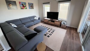 A modern living room with a grey sectional sofa, a flat-screen TV on a wooden stand, and a large window with blinds. - 14 Banksia Parade, Mallacoota (Mallacoota)