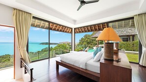 A bedroom with a large bed, a wooden nightstand, a lamp, and a view of the ocean. -  Jasmine Waterslide Villa (Koh Samui)