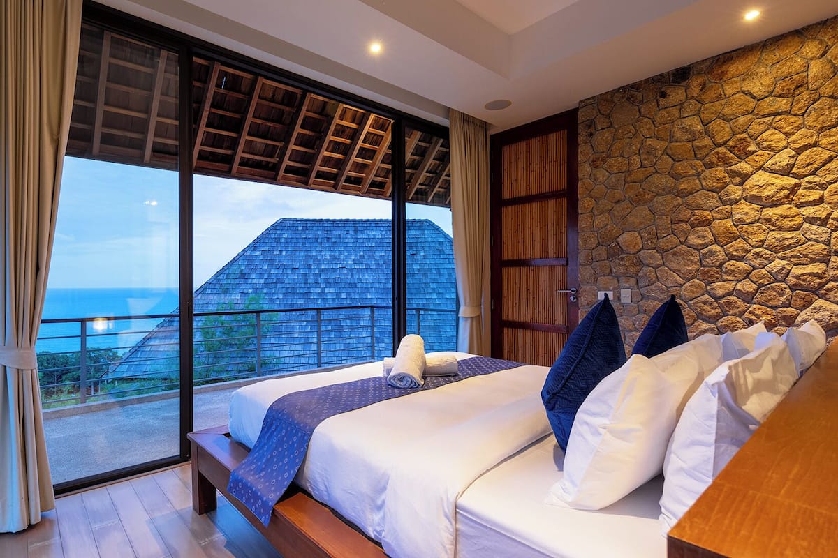A bedroom with a large window, a stone wall, and a view of a building.