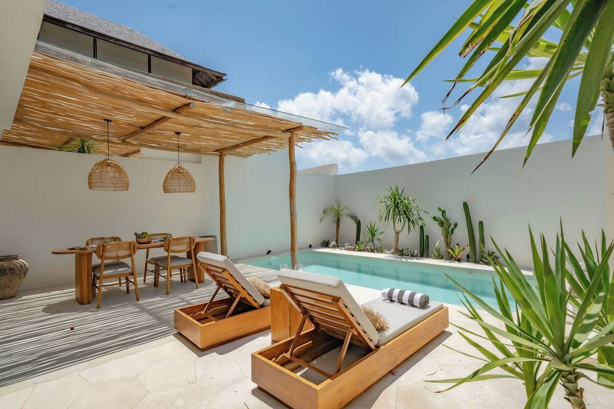 A poolside area with wooden lounge chairs, a dining table with chairs, and a pergola overhead.