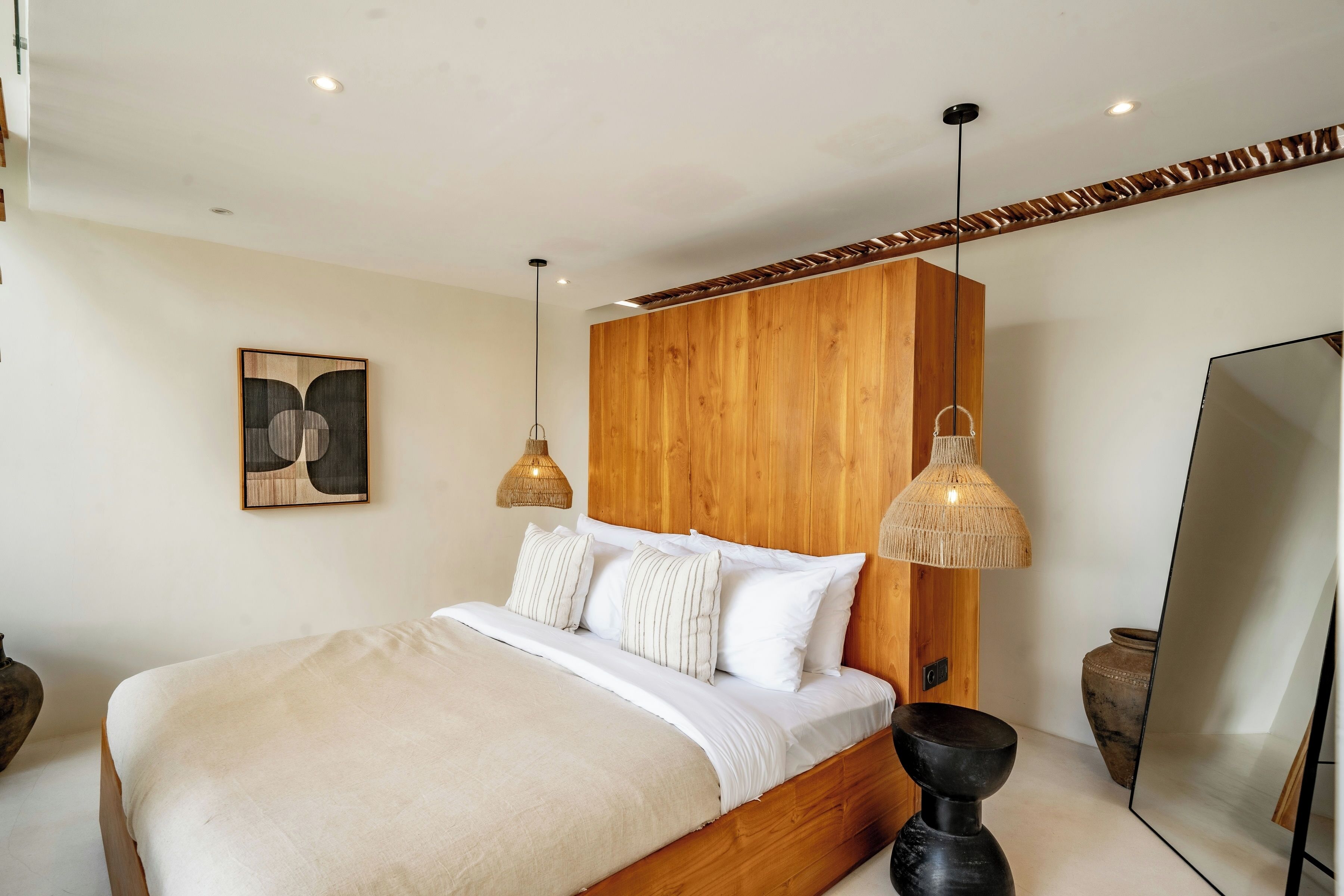 Un dormitorio moderno con cabecera de madera, una cama grande con ropa de cama blanca, dos lámparas colgantes de mimbre y un espejo grande.