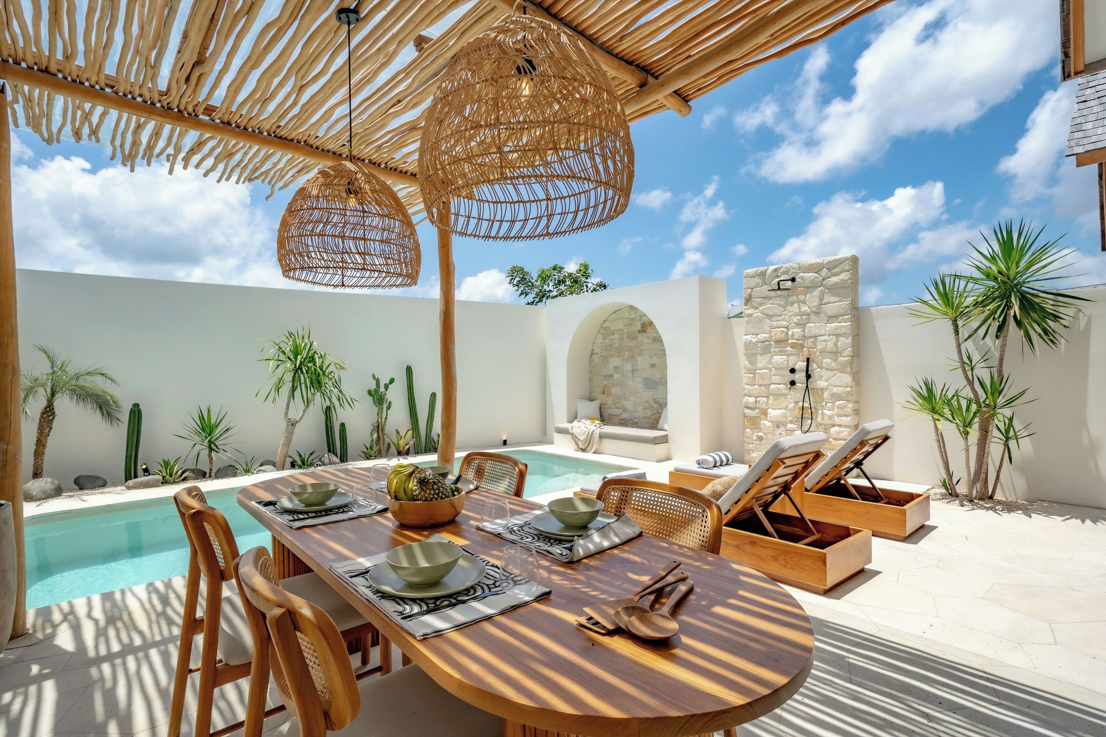 A poolside dining area with a wooden table, wicker chairs, and lounge chairs.
