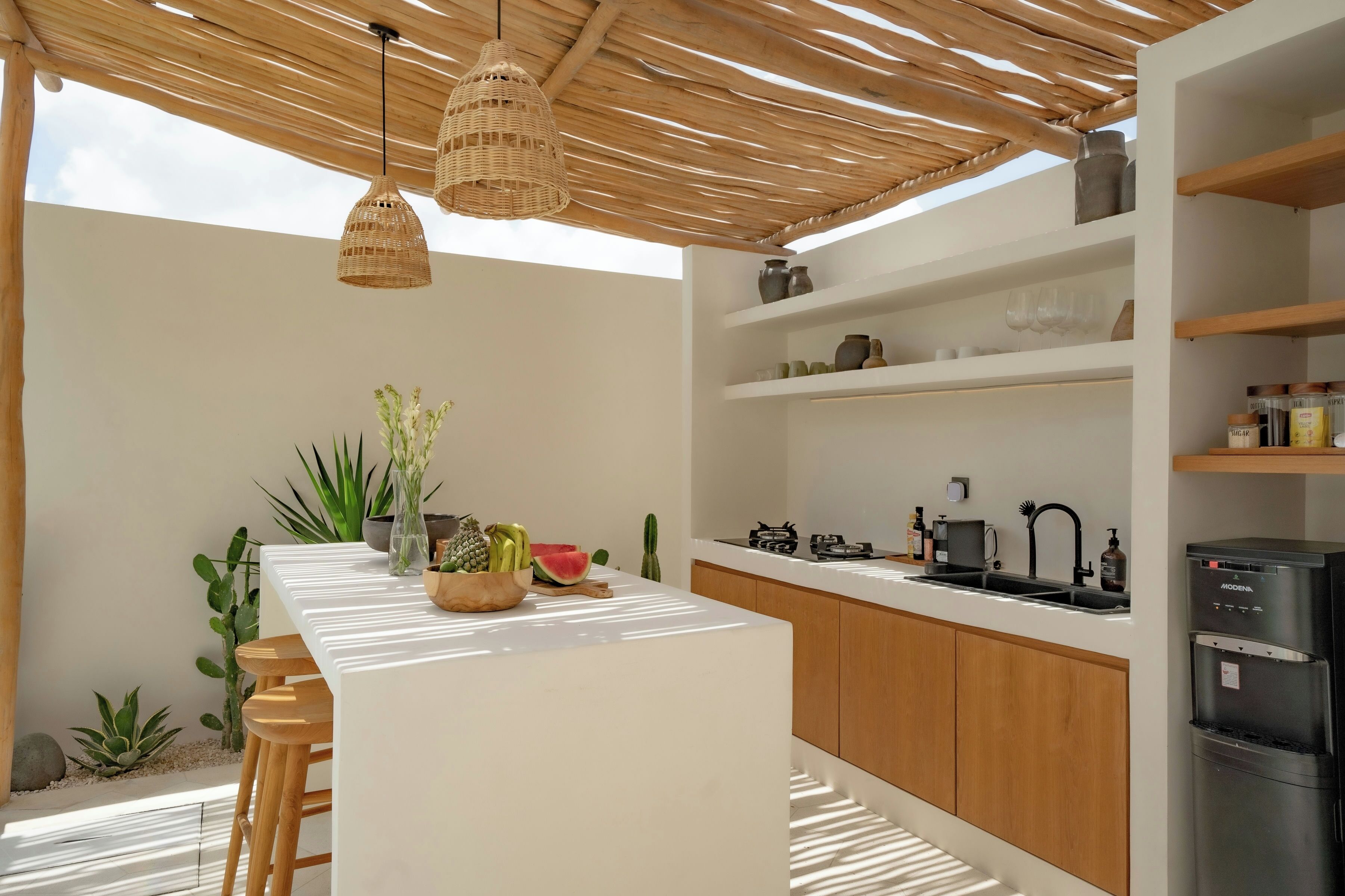 A modern kitchen with a white countertop, wooden cabinets, and a built-in refrigerator. There are wicker pendant lights, a variety of plants, and a small island with stools.