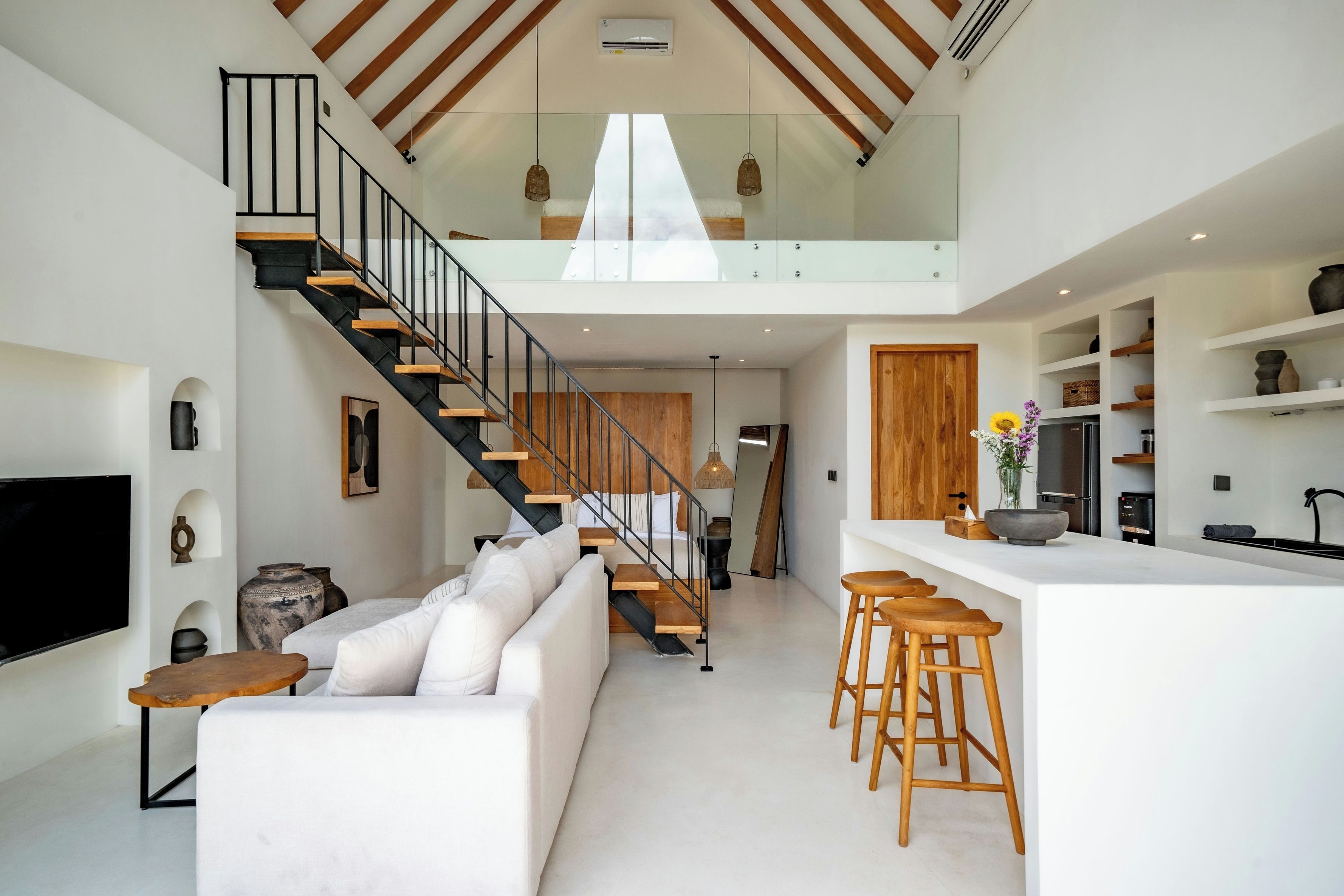 Un intérieur moderne avec un escalier menant à une mezzanine, un canapé blanc, des tabourets en bois et un coin cuisine.
