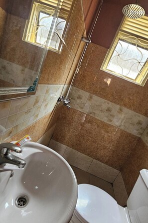 Une salle de bain avec un lavabo blanc, des toilettes et un espace douche. Il y a deux fenêtres ornées de grilles décoratives, et les murs sont recouverts de carreaux à motifs bruns.