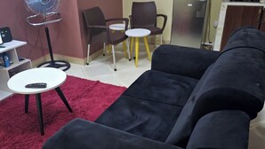Una sala de estar con un sofá negro, dos mesas redondas blancas, un tapete rojo y un ventilador de pie.