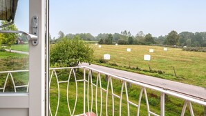 Uitzicht vanuit een raam met een metalen hekwerk, een groen veld met hooibalen, en een rode woning in de verte.