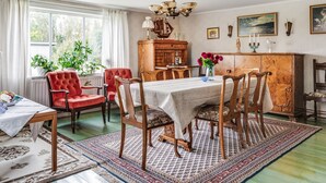 Een eetkamer met een houten tafel gedekt voor zes personen, omringd door rode fauteuils. Tegen de muur staat een houten kast met een groene bovenkant, en boven de tafel hangt een kroonluchter.