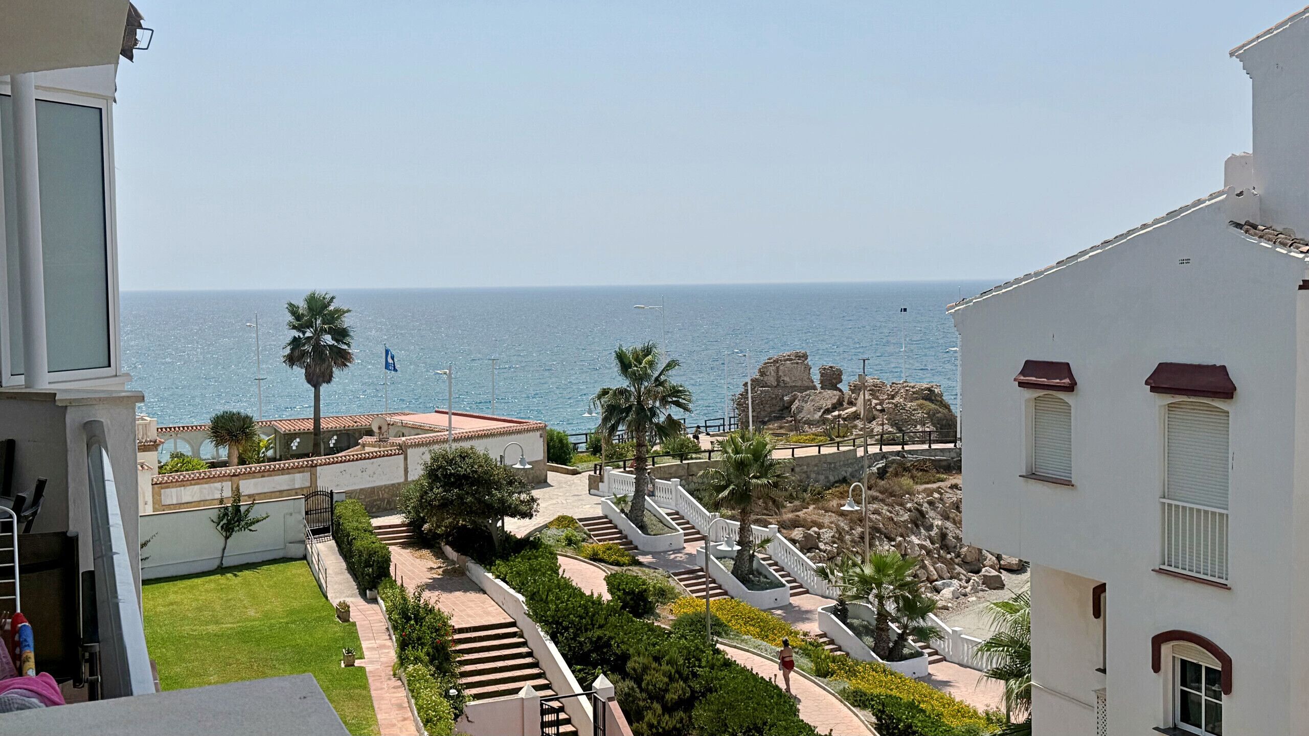 A coastal view with a clear sky, a pathway leading to a building with a white facade, and a palm tree in the foreground.