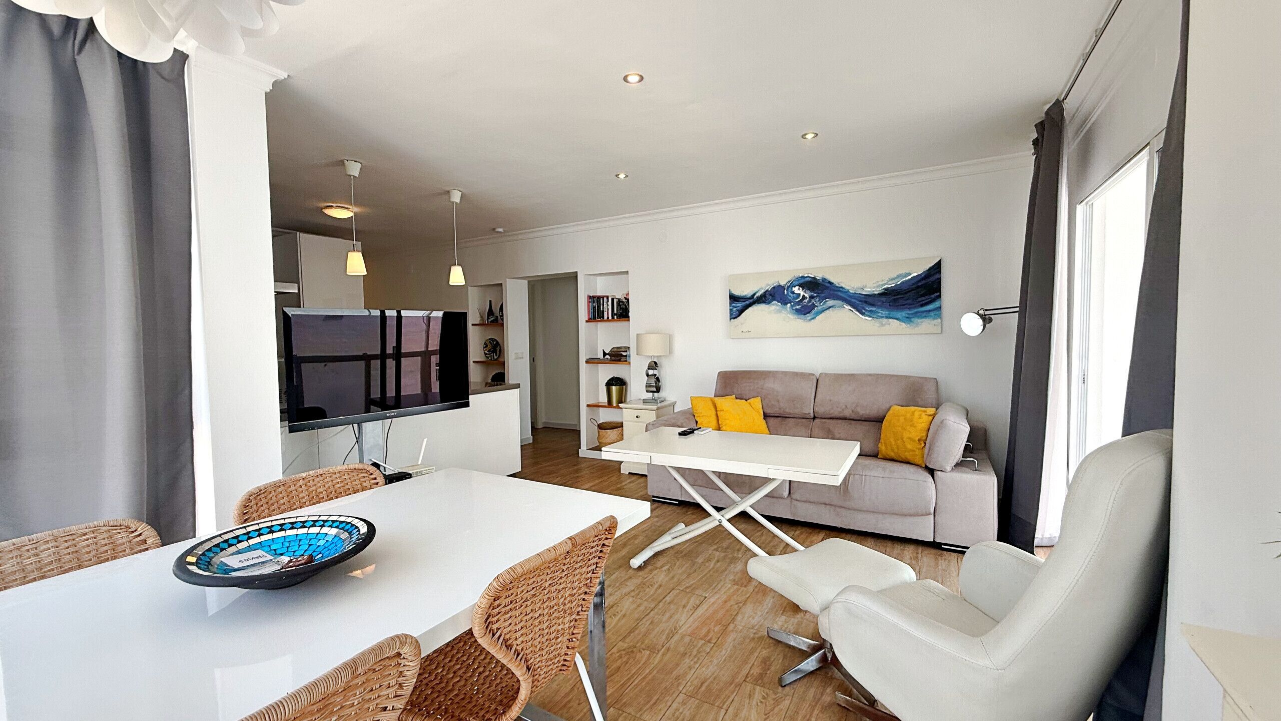 Una sala de estar moderna con sofá, mesa de comedor y sillas. Se aprecia una cocina al fondo con armarios y un refrigerador.
