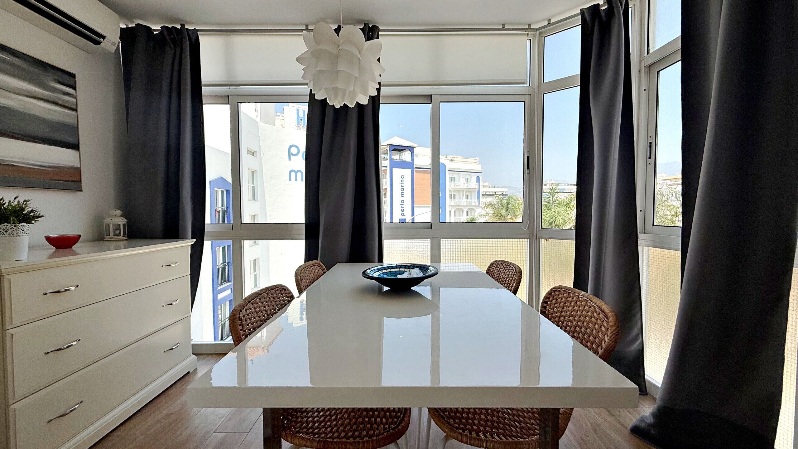 A modern dining area with a white table, wicker chairs, a central bowl, and a view of buildings through large windows.