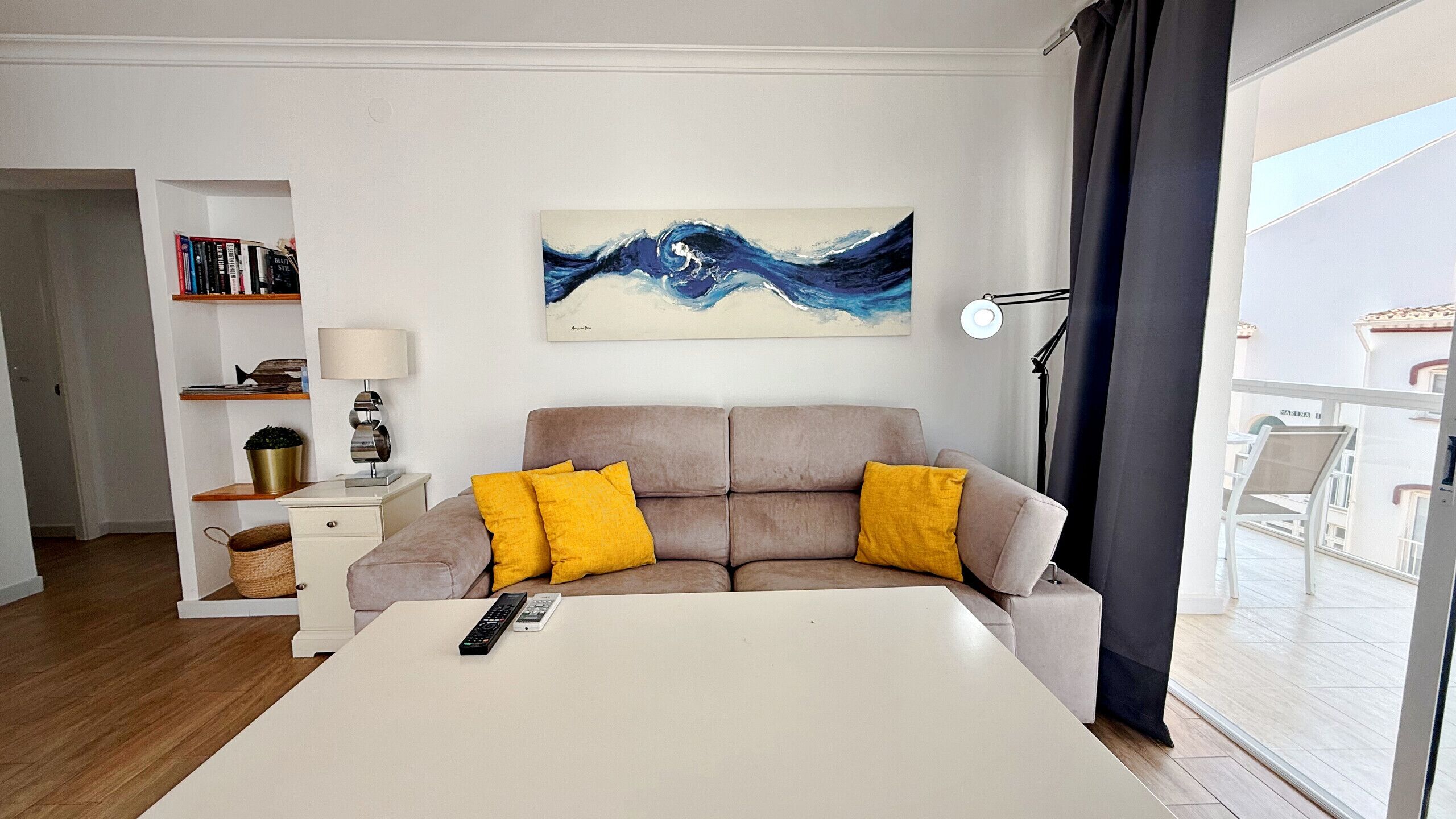 A modern bedroom with a large bed, a sofa with yellow cushions, a painting on the wall, and a view of the outside.