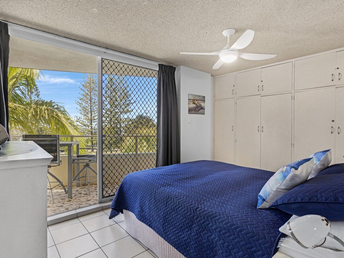 Ein Schlafzimmer mit Bett, Deckenventilator, Blick durch eine Schiebetür auf einen Außenbereich mit Tisch und Stühlen sowie einem weißen Kleiderschrank.