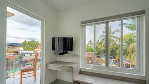 A balcony with a wooden chair, a TV mounted on the wall, and a view of palm trees and buildings. - Grace Regency (Kanayannur)