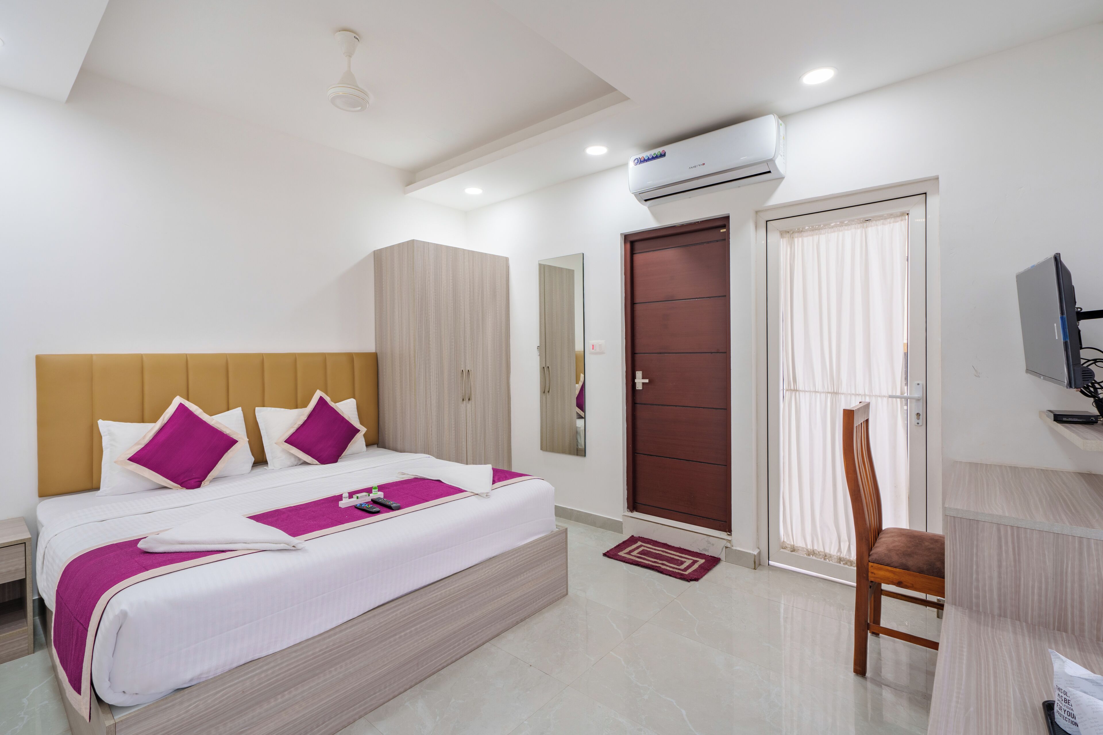 A hotel room with a bed, a desk, a chair, a wardrobe, a mirror, an air conditioning unit, and a television.