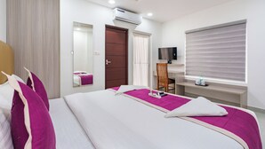 A hotel room with a bed, a desk, a chair, a TV, and an air conditioning unit. - Grace Regency (Kanayannur)