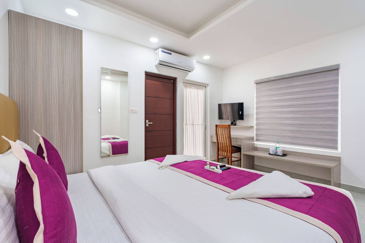 A hotel room with a bed, a desk, a chair, a TV, and an air conditioning unit.