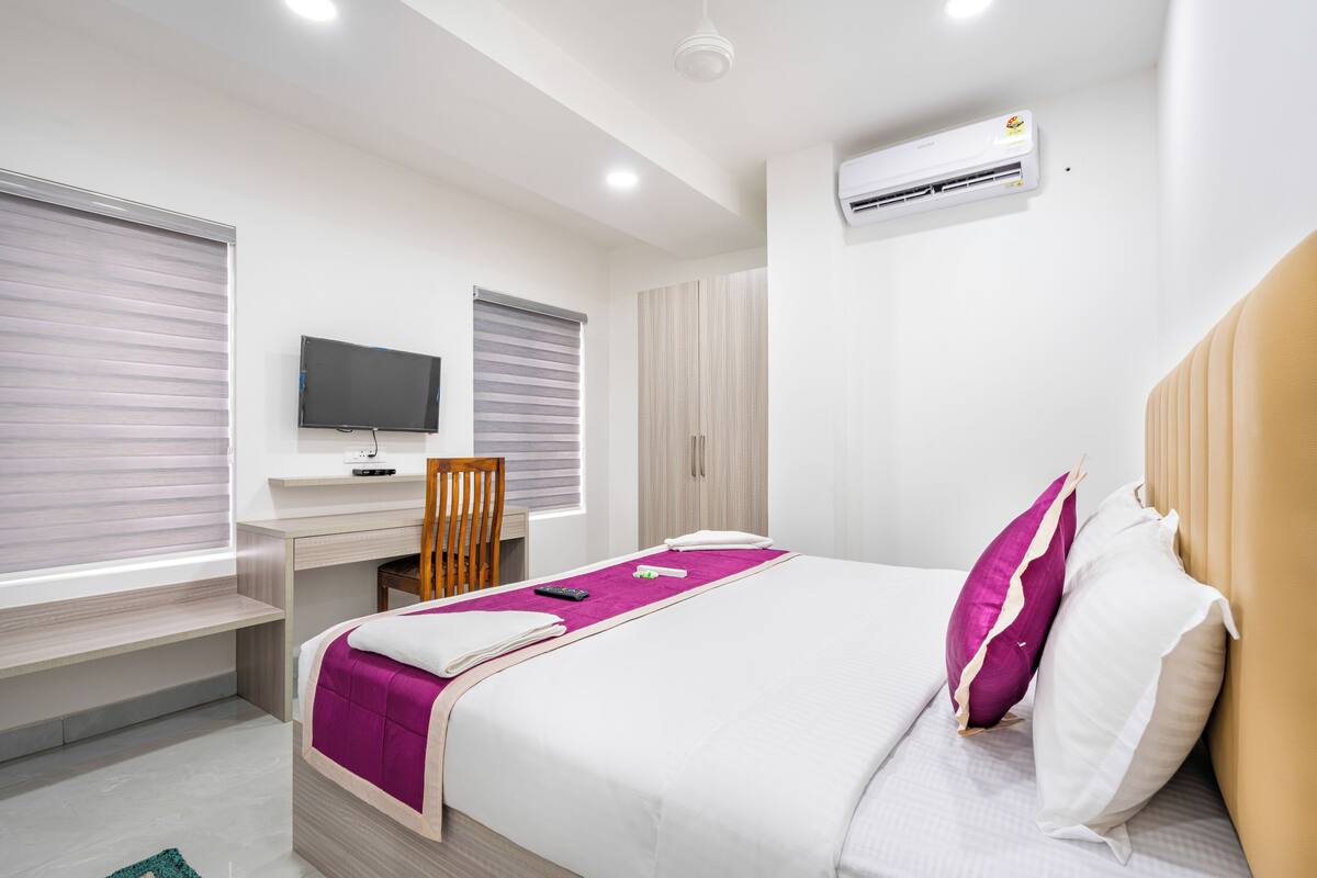 A modern hotel room with a bed, a desk, a chair, and an air conditioning unit.