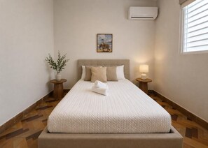 Une chambre d’hôtel bien rangée, avec un lit, une petite table sur laquelle se trouve une lampe, une plante en pot et un tableau accroché au mur.