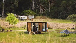 A modern, single-story cabin with a small porch, situated in a grassy field with a solar panel and a tree nearby.