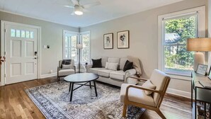 A living room with a beige sofa, a round coffee table, a wooden chair, and a lamp on a side table. There are two framed pictures on the wall and a ceiling fan. - Sweet Sage Cottage in Old Town Close to the Square (Georgetown)