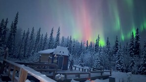 Lumipeitteinen puinen terassi, jonka yhteydessä on pieni rakennus. Terassin ympärillä on lumikuormaisia puita, ja sieltä avautuu näkymä revontuliin.