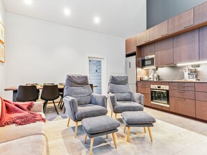 A modern living room with grey armchairs and ottomans, a dining area with a table and chairs, and a kitchen with wooden cabinets and appliances.