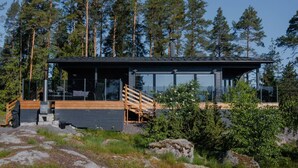 Une cabane moderne dotée d’une terrasse en bois, de grandes fenêtres et de fondations en pierre, située au cœur d’une forêt.