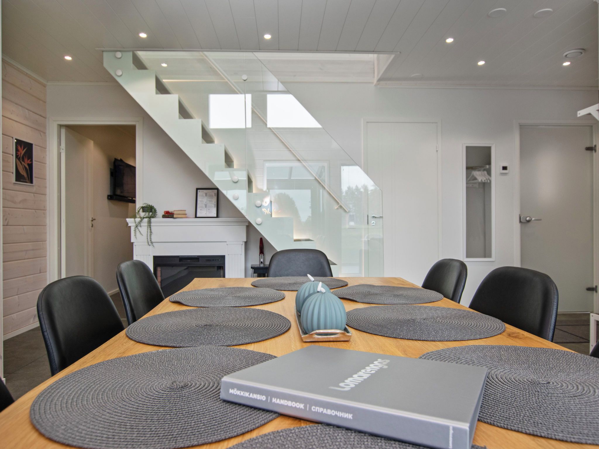 A modern dining area with a wooden table, grey placemats, and a book titled 'Möskikansio | Handbook | Sprawočnik'.