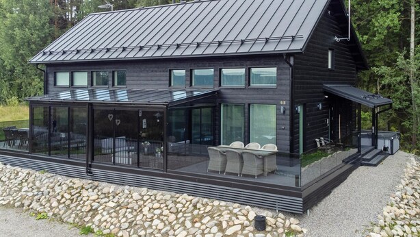 A modern black cabin with a glass-enclosed patio and a metal roof, surrounded by a gravel path and trees.