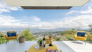 A modern room with a panoramic city view, featuring two yellow armchairs, a magazine, and a bottle of wine on a clear glass table.