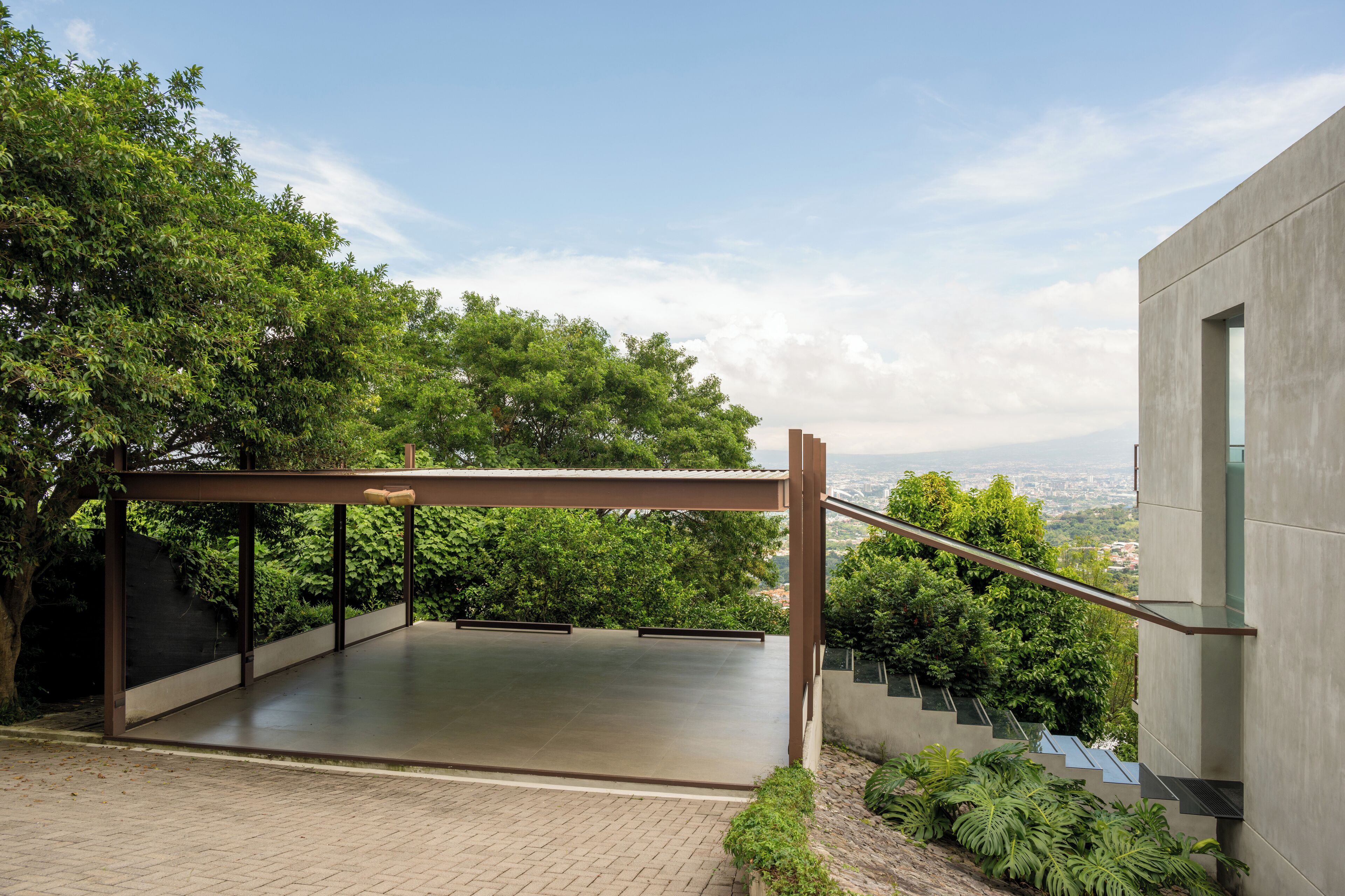 Une terrasse extérieure moderne, dotée d’une pergola en métal, entourée d’une végétation luxuriante et offrant en arrière-plan une vue sur le paysage urbain.