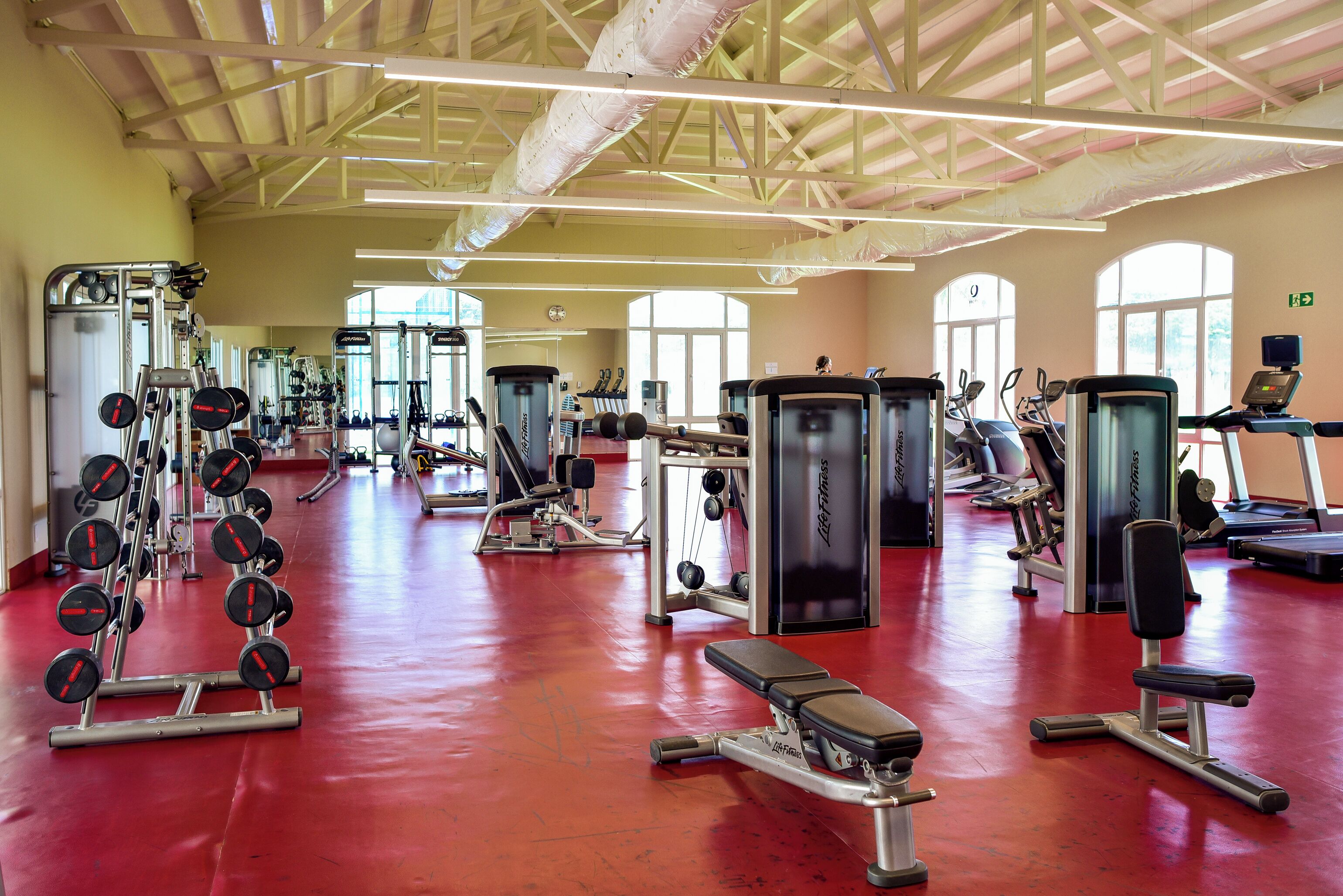 A spacious gym with various exercise equipment, large windows, and a red floor.