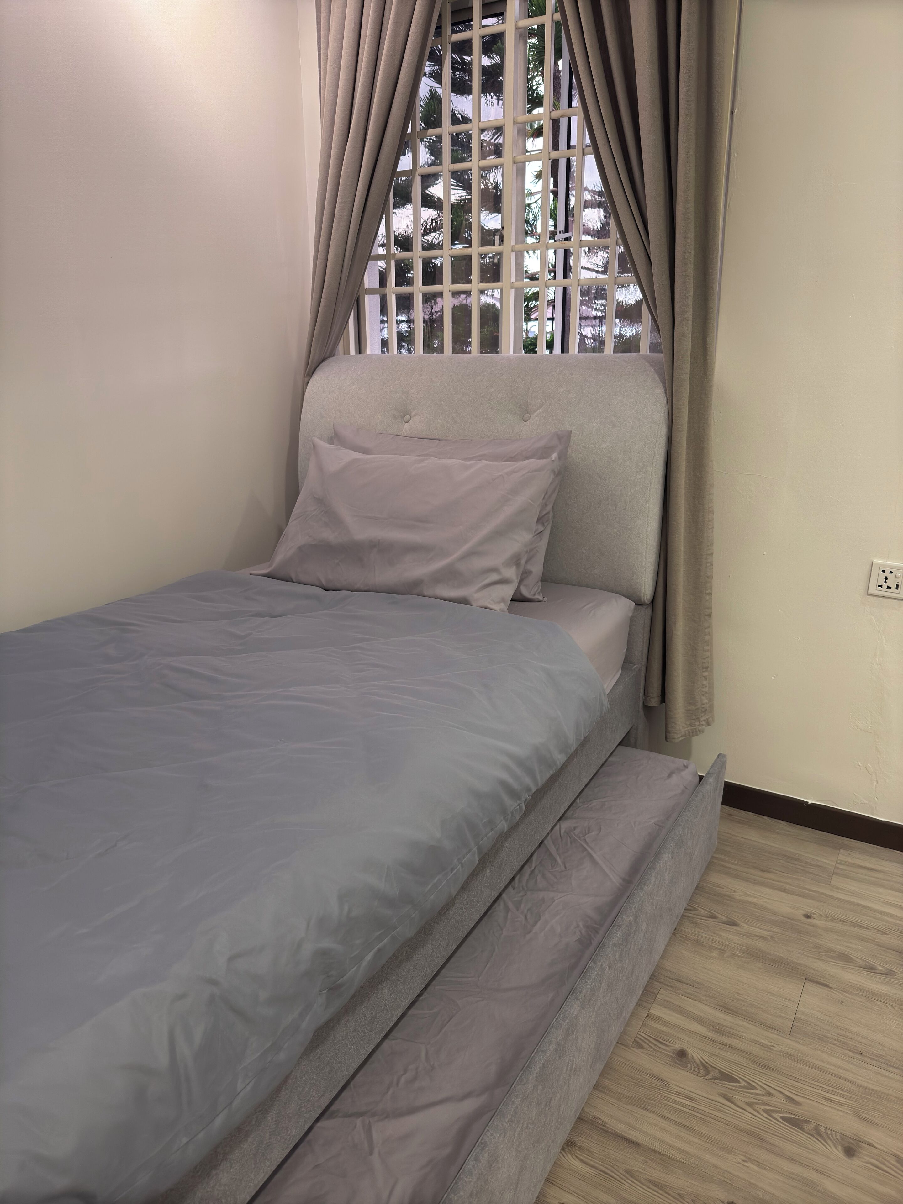 Una cama individual con colchón y almohadas grises, una ventana con cortinas y un interruptor de luz en la pared.