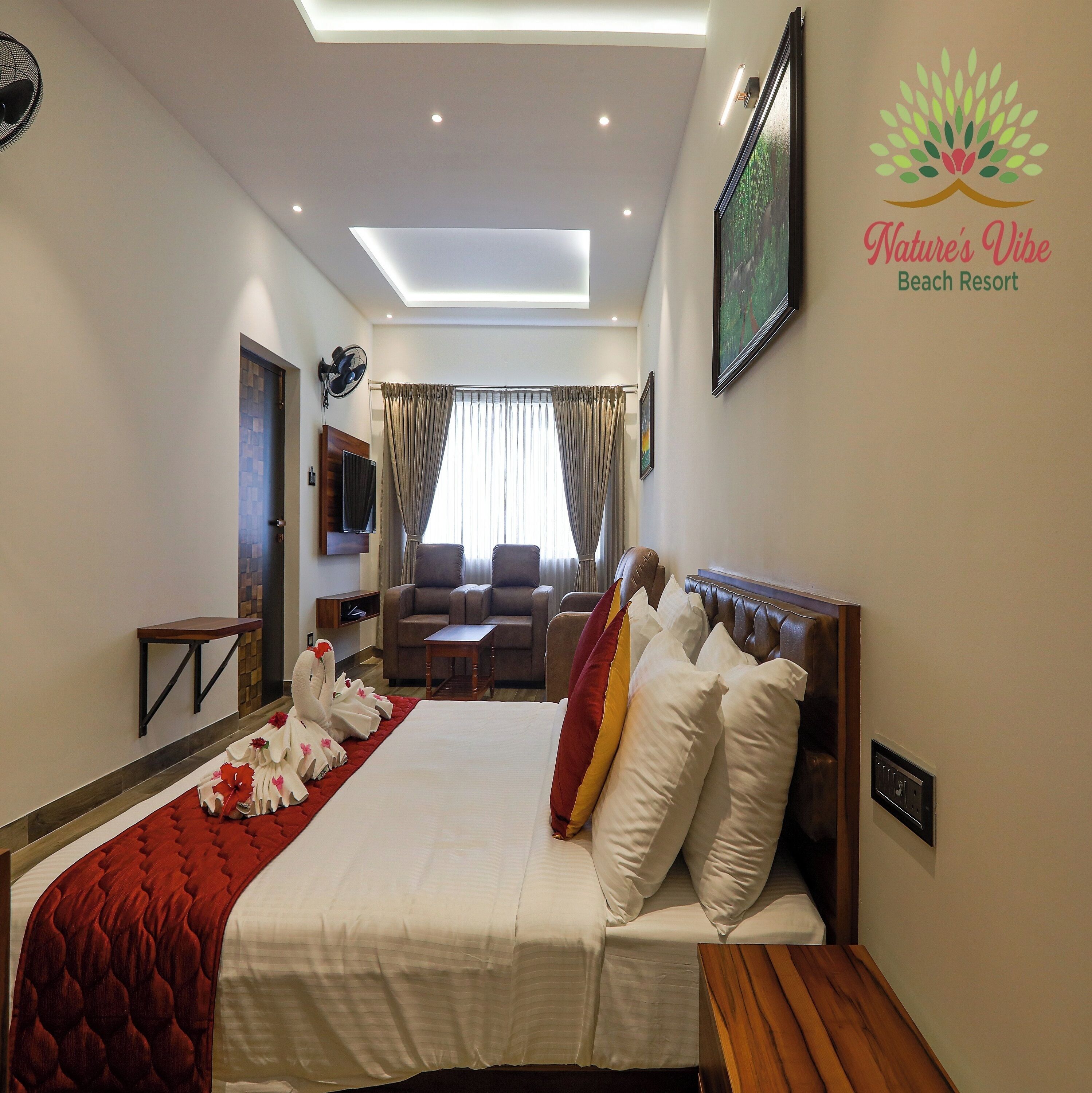 A hotel room with a bed, a TV, a fan, a window with curtains, and a wall-mounted artwork.