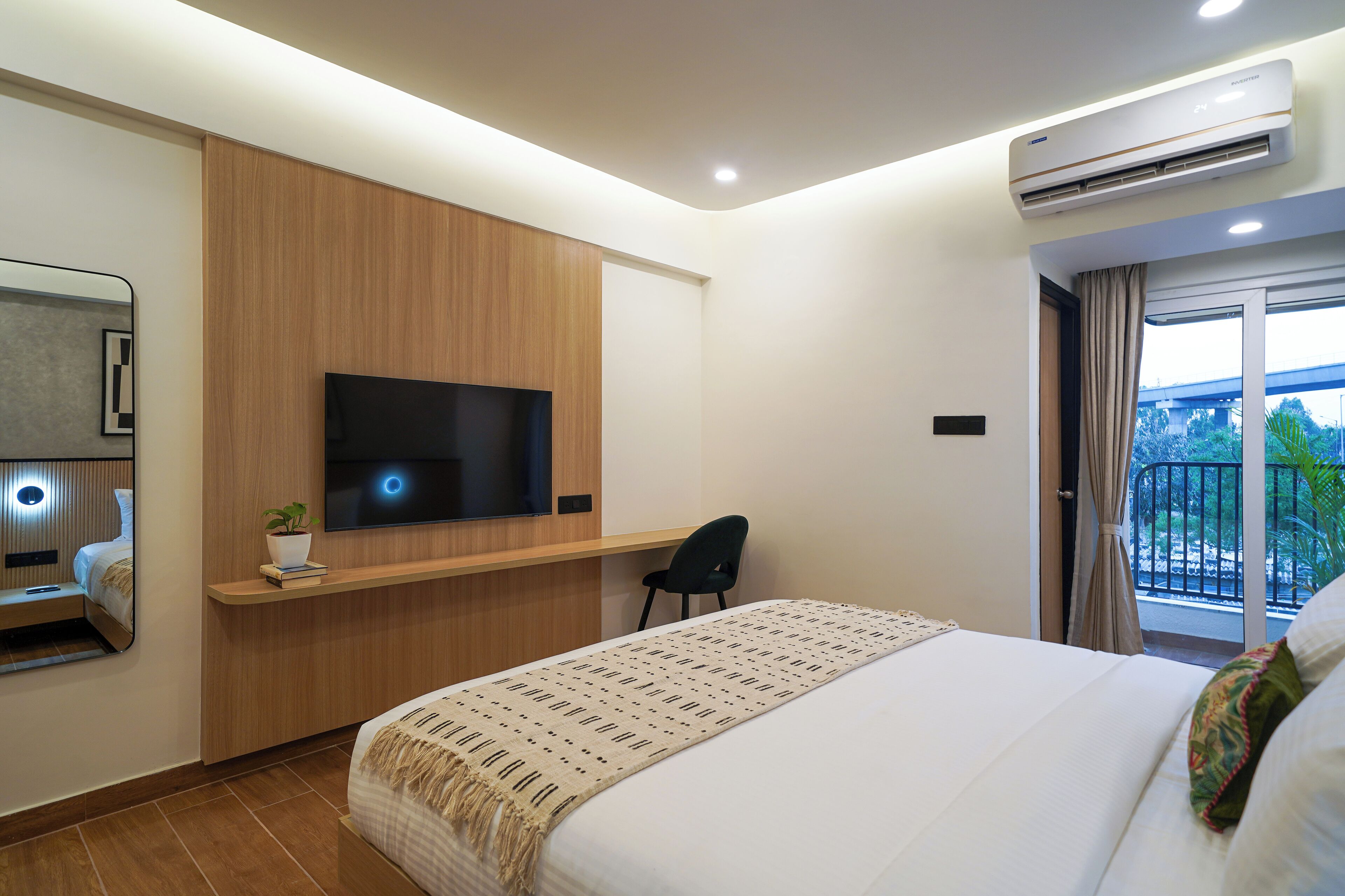 A hotel room with a bed, a TV mounted on a wooden panel, a desk with a chair, and a balcony with a view.