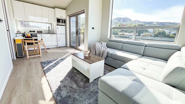 A modern kitchen with white cabinets, a dining area with a table and chairs, a grey sofa, and a large window offering a view of a green landscape. - Modern Contemporary Single Detach Home (Wellington)
