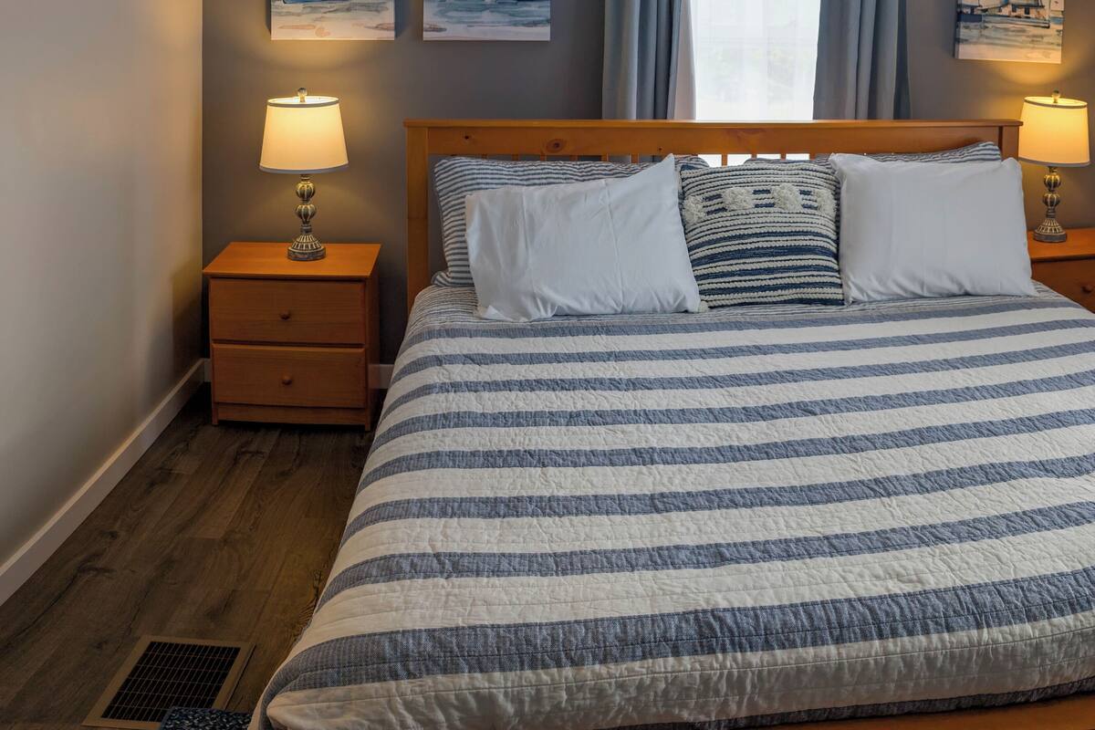 A bedroom with a bed, two bedside tables with lamps, a window with curtains, and three wall-mounted paintings of sailboats.