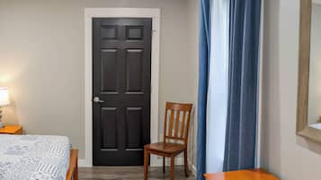 Quarto com cama, cómoda de madeira, cadeira, porta e janela com cortinas.
