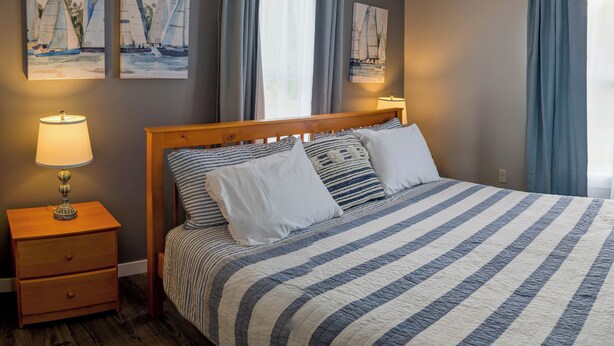 A neatly made bed with striped bedding, two bedside tables with lamps, and two paintings of sailboats on the wall. - Newly Renovated Acadia Apartment Maple Street (Ellsworth)