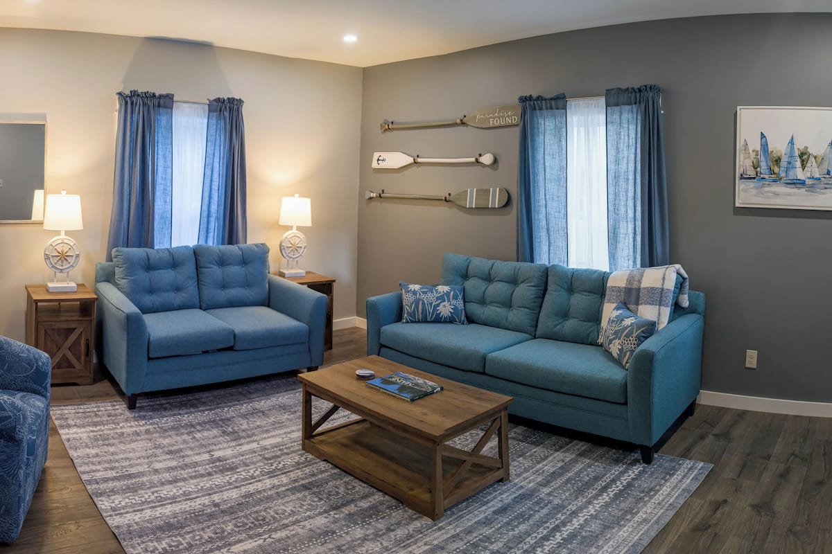 A living room with two blue sofas, a coffee table, and a rug. There are wall-mounted lamps and a painting of sailboats.