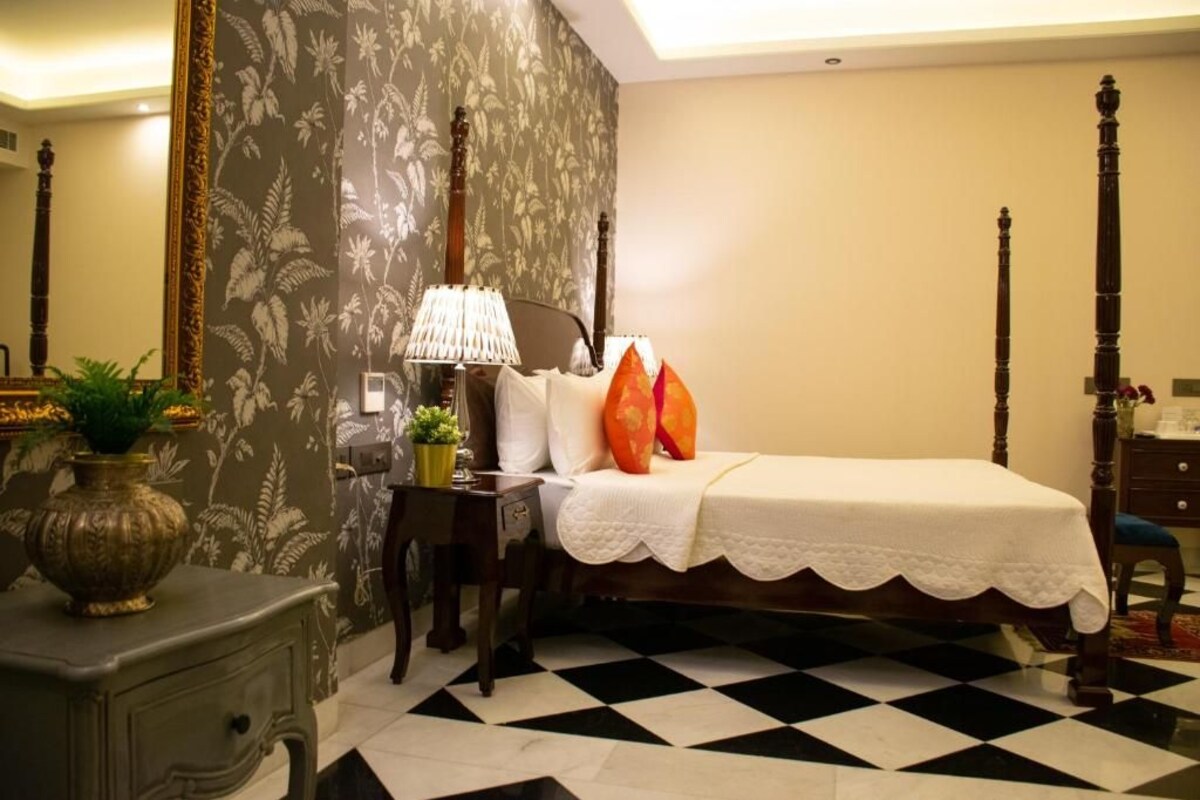 A bedroom with a checkered floor, a bed with white linens, a nightstand with a lamp, and a floral wallpaper.